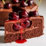 A slice of homemade chocolate cherry cake with rich chocolate layers, creamy frosting, and a topping of fresh cherries and cherry sauce, dripping onto a white plate.