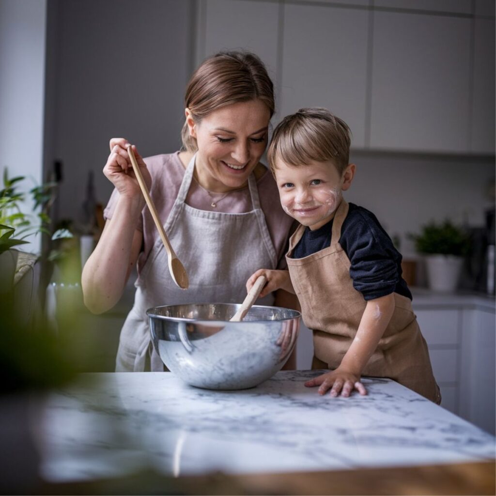 A candid iPhone photo of food blogger Hannah and her 6-year-old son Leo in their bright home kitchen. Hannah is laughing while holding a wooden spoon, wearing a beige linen apron, as Leo stands on a kitchen stool with flour dusted playfully across his cheeks. They're sharing a joyful moment while baking together at their marble countertop.