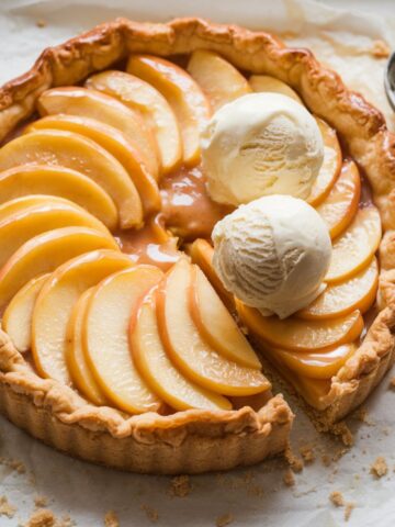 Rectangular apple tart with golden-brown puff pastry, glazed apple slices, and melting vanilla ice cream, on parchment paper with an ice cream scoop nearby