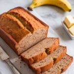 A freshly baked banana bread with two bananas loaf with a golden crust, sliced to reveal its moist, fluffy interior.