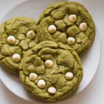 Three freshly baked matcha cookies with white chocolate chips cooling on a white plate set on a white kitchen counter.