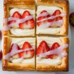 Golden and flaky strawberry puff pastry slices, topped with fresh sliced strawberries, creamy filling, and a drizzle of white icing on parchment paper.