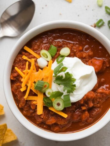 A bowl of Brooks seasoning chili topped with cheese, sour cream, and green onions on a white kitchen counter with tortilla chips scattered nearby