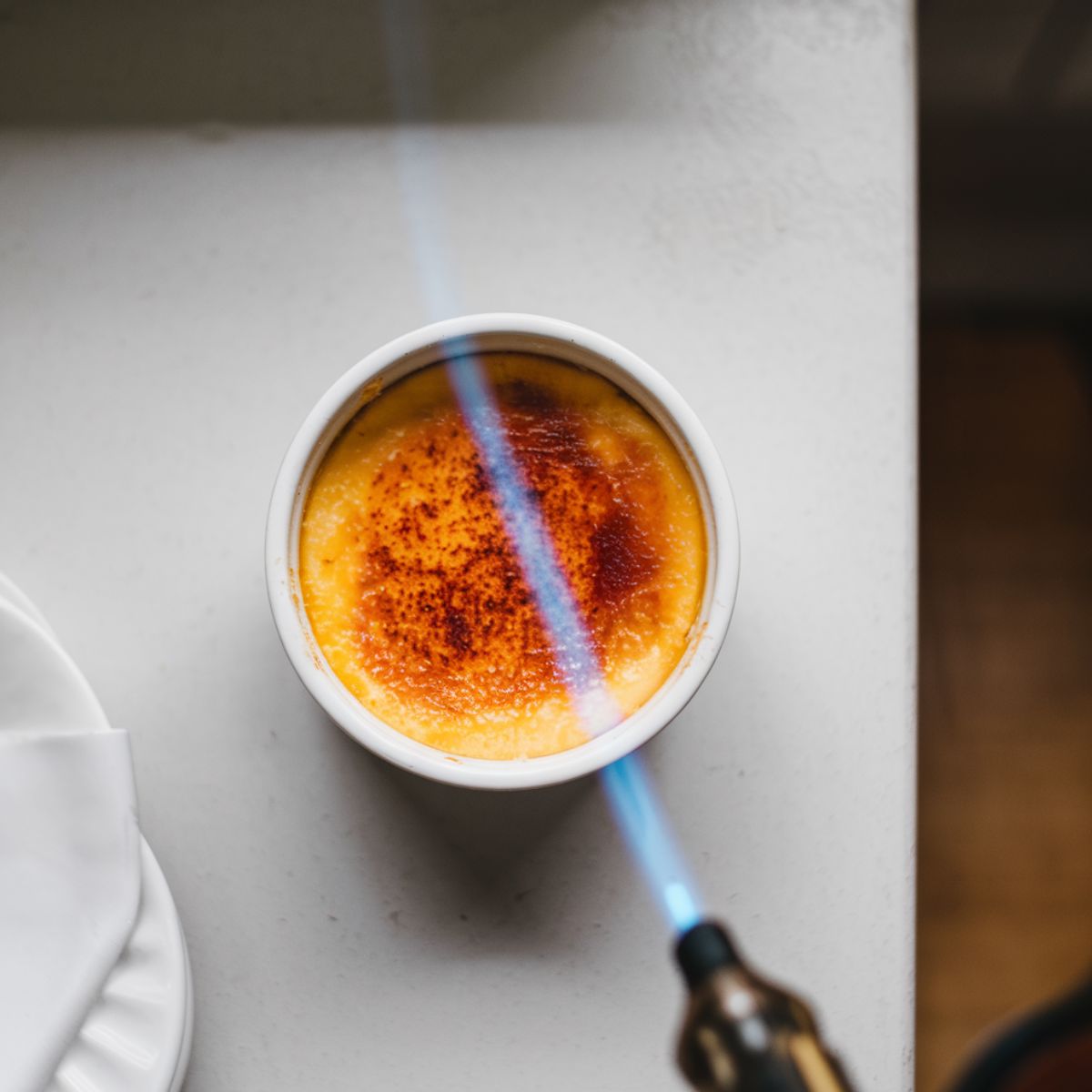 A ramekin filled with crab brulee, featuring a smooth, golden caramelized top being torched with a blue flame. The setting is minimal, with the ramekin near a plate and napkin, highlighting the process of achieving the signature crust