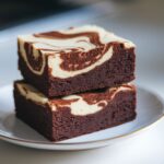 Two fudgy cream cheese brownies with a rich chocolate base and smooth marbled cream cheese swirl, stacked on a white plate, perfect for a dessert recipe.