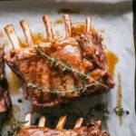 A plate of perfectly cooked lamb ribs with caramelized glaze, garnished with rosemary and thyme, on a white kitchen counter.