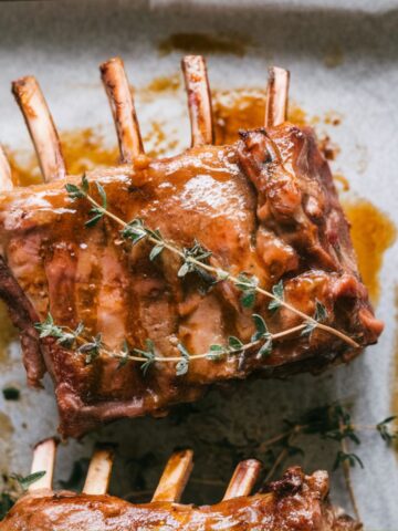 A plate of perfectly cooked lamb ribs with caramelized glaze, garnished with rosemary and thyme, on a white kitchen counter.