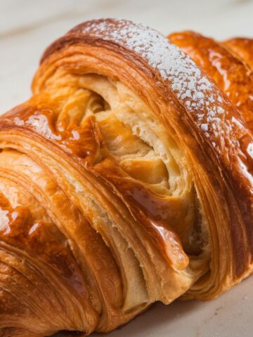 Close-up of a golden-brown Swiss gipfeli (croissant) with flaky, layered pastry, a glossy caramelized surface, and a light dusting of powdered sugar, resting on parchment pape