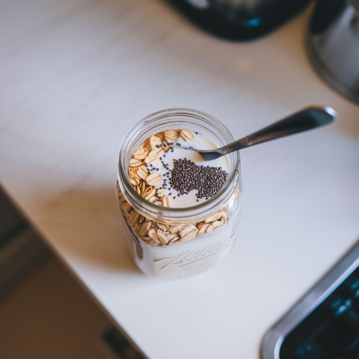 A mason jar filled with oats, protein powder, chia seeds, and milk being stirred with a spoon on a white kitchen counter.