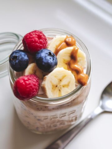 A jar of protein overnight oats topped with fresh berries, banana slices, and peanut butter on a white kitchen counter.