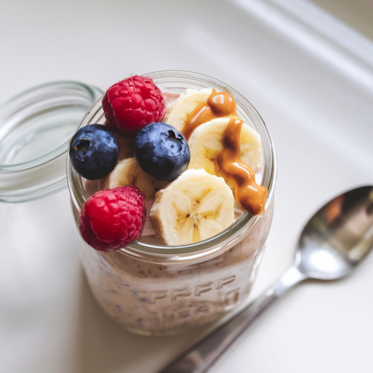 A jar of protein overnight oats topped with fresh berries, banana slices, and peanut butter on a white kitchen counter.