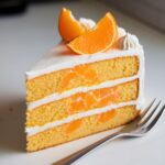 A slice of layered mandarin orange cake with fresh mandarins and whipped cream on a white plate, accompanied by a fork.