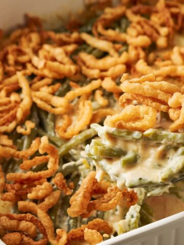 A homemade Campbell Soup green bean casserole with golden-brown crispy onions on top, sitting on a white kitchen counter. The casserole has bubbling creamy sauce around the edges, with a serving spoon holding a huge portion of the dish