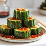 Stacked cucumber slices drizzled with a garlicky chili soy sauce, served on a ceramic plate with chopsticks on a white kitchen counter.