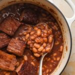 A pot of slow-cooked pork and beans recipe with tender chunks of pork and creamy beans in a rich, savory sauce, with a spoon lifting a portion.