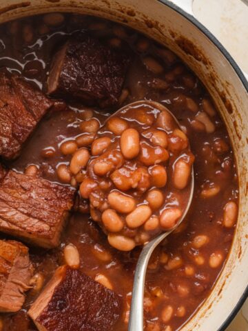 A pot of slow-cooked pork and beans recipe with tender chunks of pork and creamy beans in a rich, savory sauce, with a spoon lifting a portion.