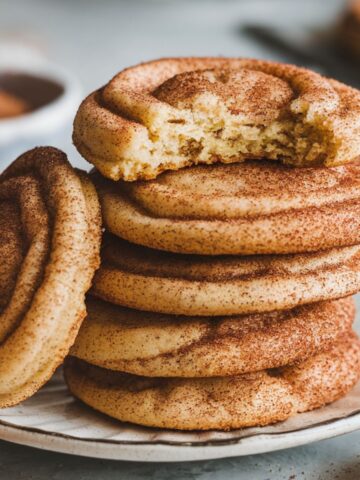 A stack of golden-brown snickerdoodle cookies with cinnamon-sugar coating on a white plate, placed on a home kitchen counter.