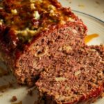 A homemade stove top stuffing meatloaf with a caramelized glaze, sliced on a white plate with crumbs scattered on the counter.