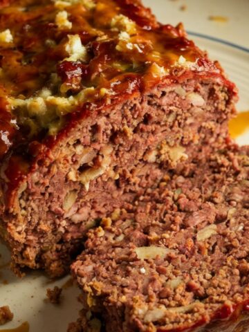 A homemade stove top stuffing meatloaf with a caramelized glaze, sliced on a white plate with crumbs scattered on the counter.