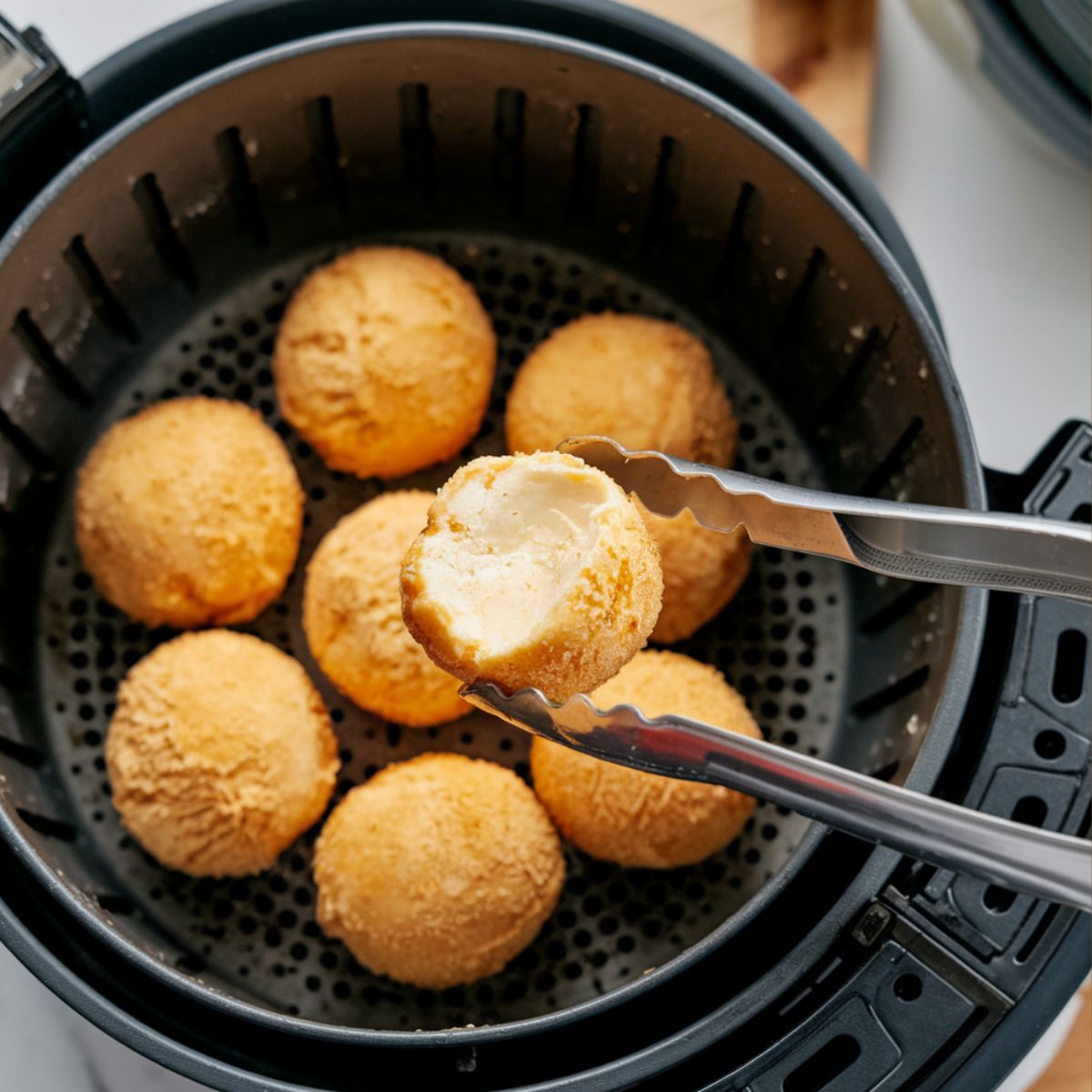 Golden, crispy air fryer fried ice cream balls inside an air fryer basket, one being lifted with tongs.