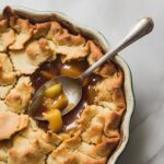 Freshly baked apple pandowdy in a white square ceramic dish with a golden, cracked crust and bubbling caramelized apple filling, with a silver spoon scooping into the dessert.