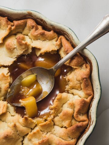 Freshly baked apple pandowdy in a white square ceramic dish with a golden, cracked crust and bubbling caramelized apple filling, with a silver spoon scooping into the dessert.