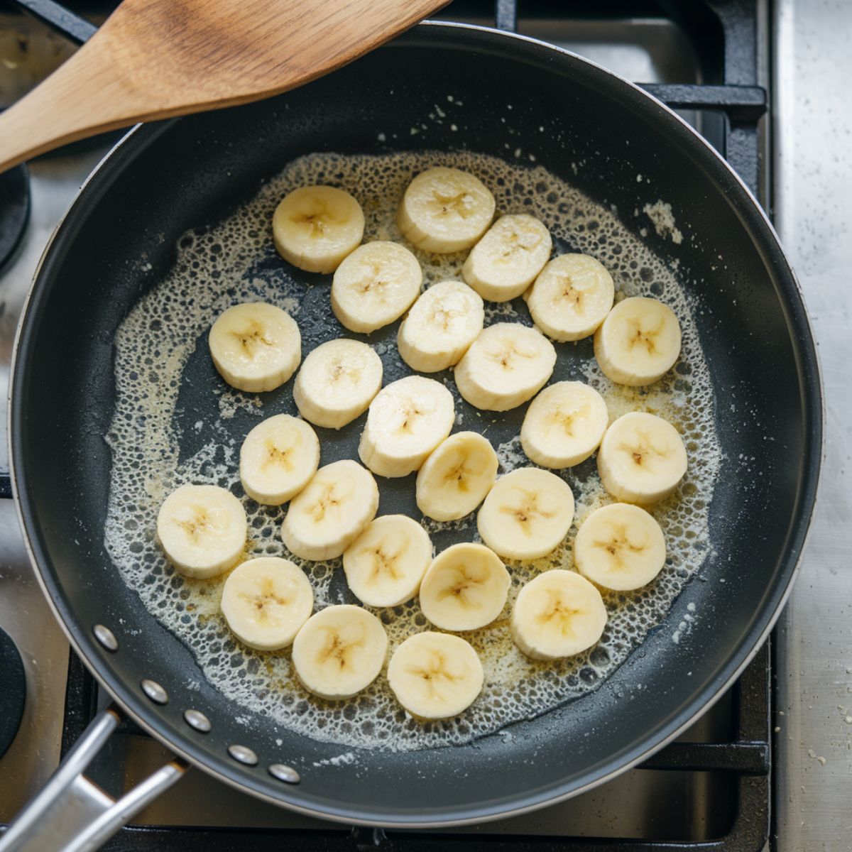 Sliced bananas sizzling in bubbling butter in a non-stick pan on a stovetop, with a wooden spatula nearby.