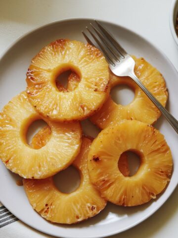 Plate of homemade caramelized pineapple rings with golden edges, a fork, and brown sugar on a white kitchen counter.