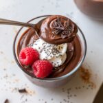 A glass bowl of homemade chocolate protein pudding topped with whipped cream, chia seeds, raspberries, and dark chocolate shavings, with a spoon lifting a creamy bite.