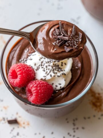 A glass bowl of homemade chocolate protein pudding topped with whipped cream, chia seeds, raspberries, and dark chocolate shavings, with a spoon lifting a creamy bite.