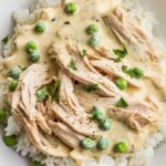 A homemade plate of creamed chicken over rice, with a thick, creamy sauce, shredded chicken, green peas, and a garnish of fresh parsley.