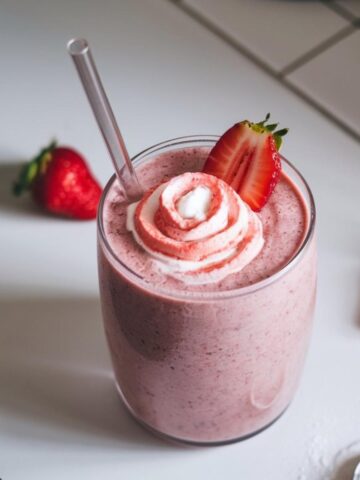 A homemade Hailey Bieber smoothie in a clear glass, topped with coconut cream and fresh strawberries, on a white kitchen counter."