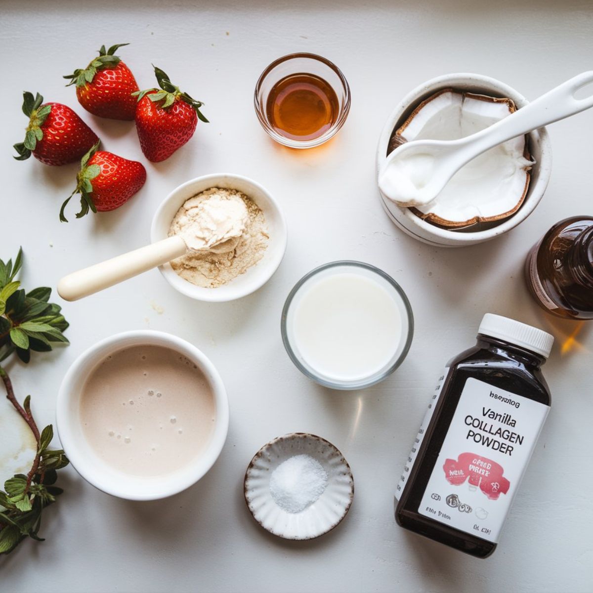 A top-down view of hailey bieber smoothie recipe ingredients including fresh strawberries, collagen powder, coconut cream, almond milk, maple syrup, and sea salt on a white kitchen counter.