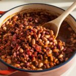 A pot of homemade calico beans with kidney beans, navy beans, and bacon in a thick sauce. A wooden spoon stirs the mixture under warm natural light.