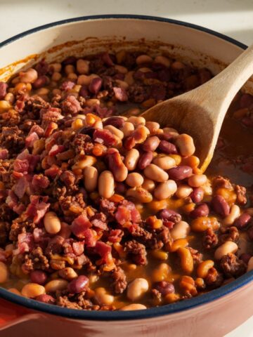 A pot of homemade calico beans with kidney beans, navy beans, and bacon in a thick sauce. A wooden spoon stirs the mixture under warm natural light.