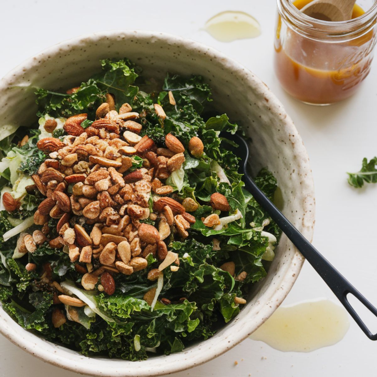 A homemade Chick-fil-A Kale Crunch Salad in a white ceramic bowl, topped with almonds, sunflower seeds, and apple chips, next to a mason jar of dressing on a white kitchen counter.