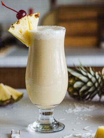 A homemade Coco Lopez Piña Colada in a glass, topped with pineapple and cherry, sitting on a white kitchen counter.