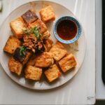 A plate of crispy, golden-brown homemade tauhu with dipping sauce, garnished with fresh herbs and crispy shallots, placed on a white kitchen counter