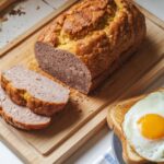 Sliced homemade scrapple loaf on a wooden board, showing crispy edges and soft interior, served with eggs and toast on a white kitchen counter.