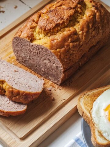 Sliced homemade scrapple loaf on a wooden board, showing crispy edges and soft interior, served with eggs and toast on a white kitchen counter.