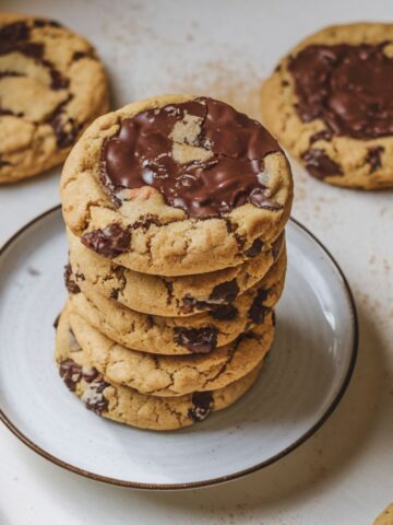 A stack of freshly baked Jacques Torres chocolate chip cookies on a white plate, surrounded by more cookies and scattered chocolate chunks on a white countertop.