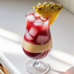 A homemade Malibu Bay Breeze cocktail with ice, layered cranberry and pineapple juice, and a pineapple wedge garnish, sitting on a white kitchen counter.