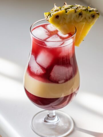 A homemade Malibu Bay Breeze cocktail with ice, layered cranberry and pineapple juice, and a pineapple wedge garnish, sitting on a white kitchen counter.