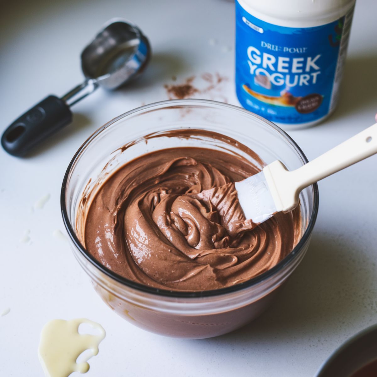 A thick chocolate protein pudding mixture being stirred in a clear bowl with a spatula, with ingredients scattered on a white kitchen counter.