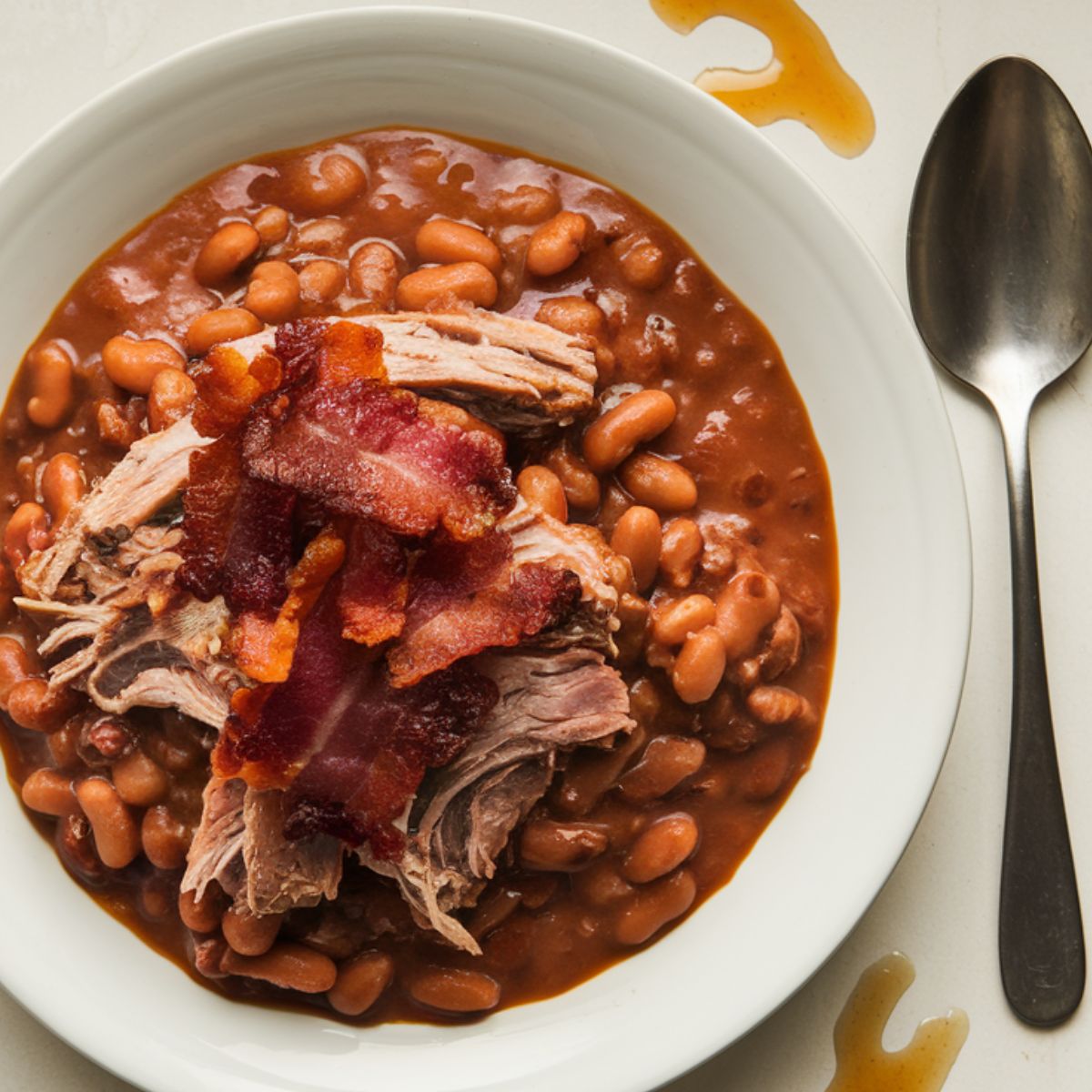 A plate of homemade pork and beans, featuring tender shredded pork topped with crispy bacon, served in a rich, savory sauce.