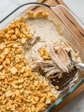 Creamy poppy seed chicken casserole with a golden, crunchy cracker topping in a glass baking dish. A spoon scoops out a portion, revealing tender shredded chicken.