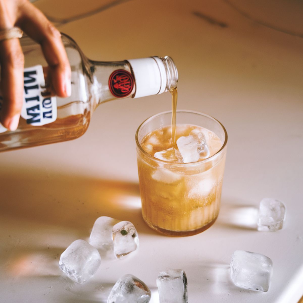 A hand pouring Malibu coconut rum into a glass filled with ice on a white kitchen counter, with a few scattered ice cubes around.