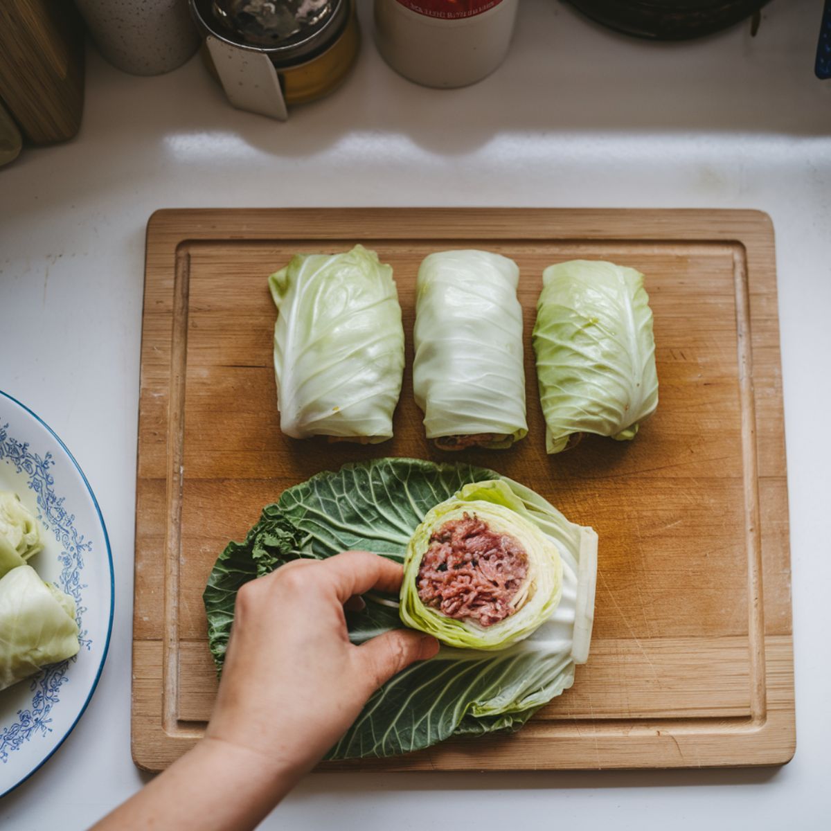 A hand folding a cabbage leaf around a meat and rice filling on a white cutting board, with pre-rolled golumpki nearby.