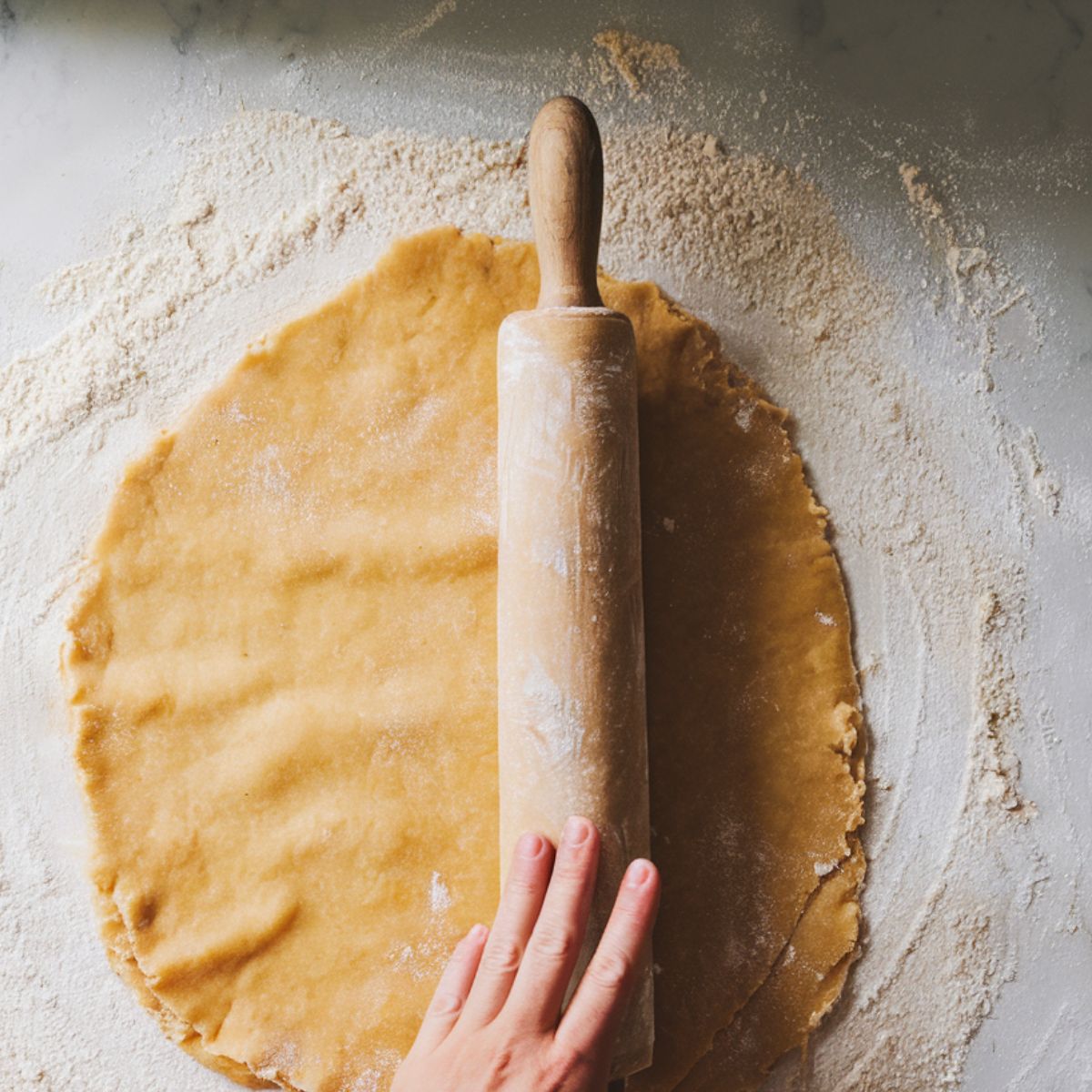 Homemade pastry dough being rolled out on a floured kitchen counter with a rolling pin, showing a rustic and uneven texture.