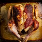 A top-down view of a partially cooked rotisserie chicken on a roasting tray, with crispy golden-brown skin developing, surrounded by rendered juices and seasoning.